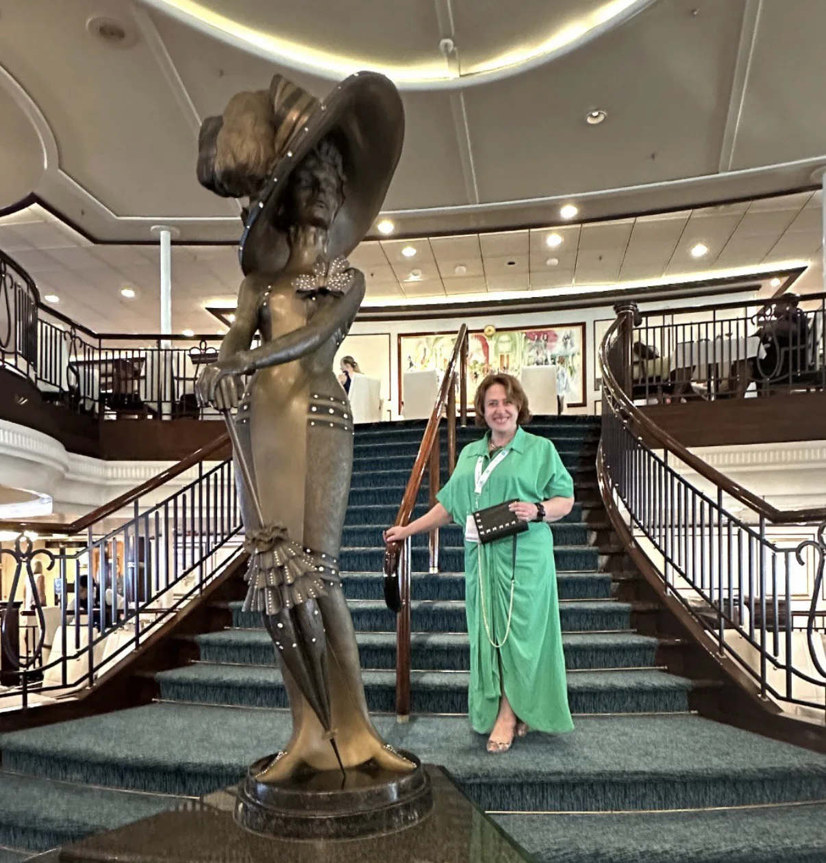 Женщина в зеленом платье стоит на лестнице рядом со статуей в интерьере здания