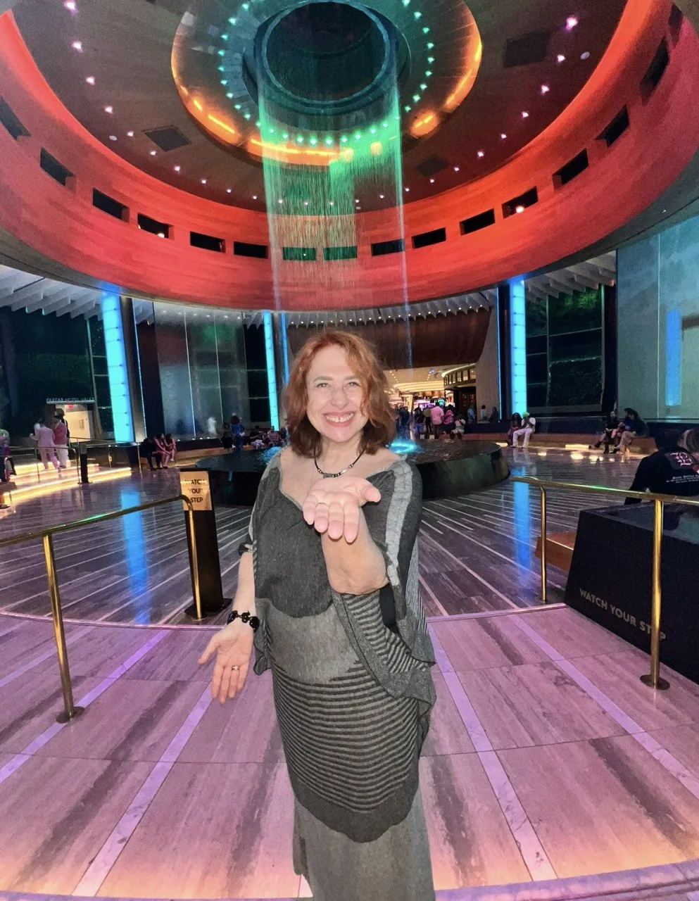 Женщина в сером платье протягивает руку в просторном холле с круглой световой инсталляцией на потолке